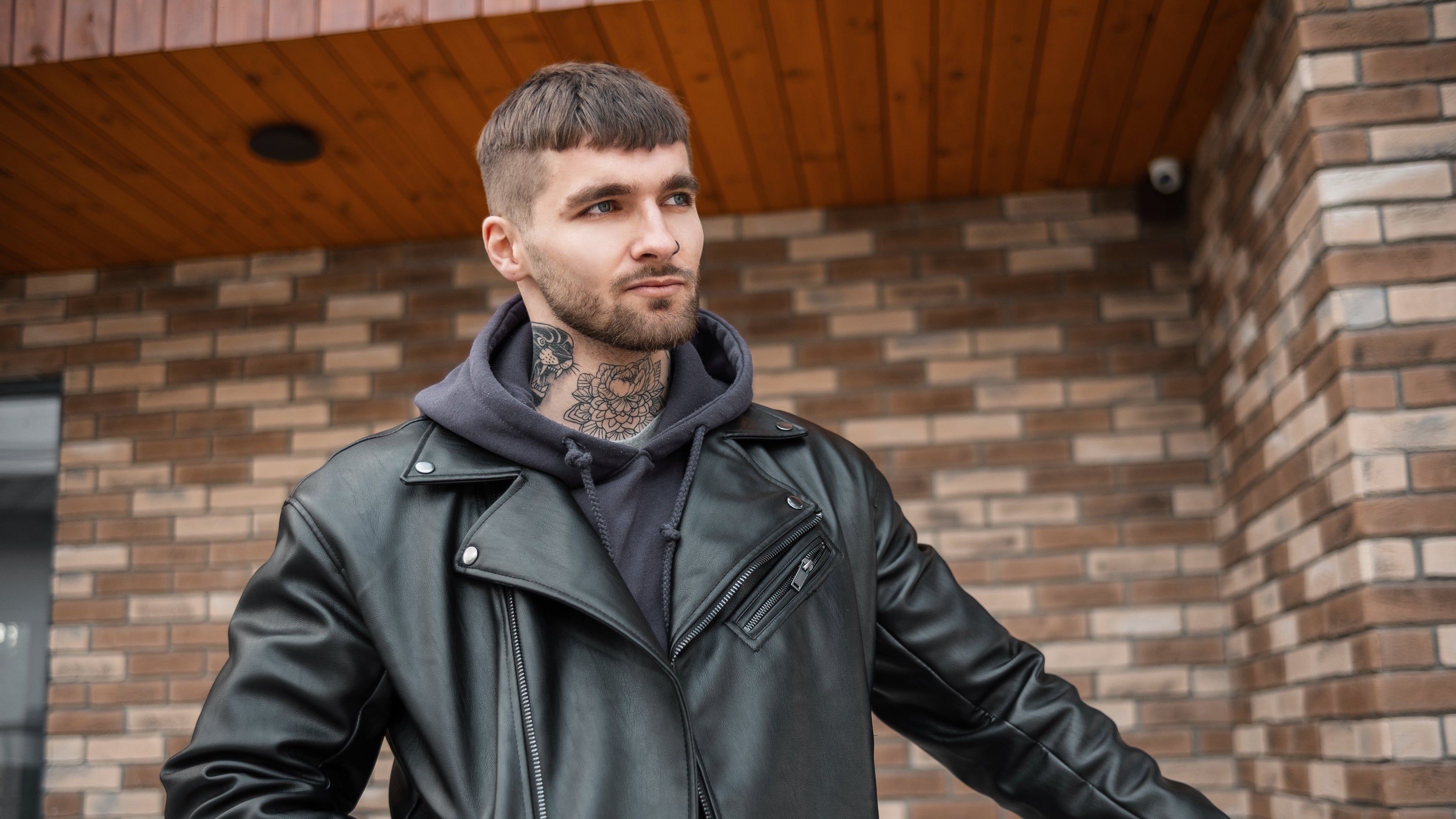 Man wearing a black leather jacket and hoodie standing in front of a brick wall.